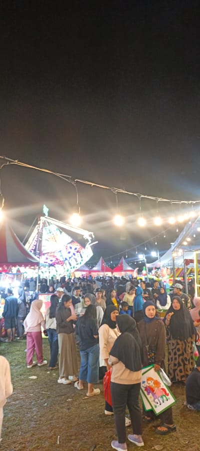 The image depicts an outdoor nighttime scene at a local event in Sinar Semendo, Indonesia. A large crowd of people are gathered, illuminated by strings of bare light bulbs strung across the dark sky. In the background, several tents with red and white striped awnings are visible. A prominent colorful structure, possibly a small ride or attraction, is illuminated with vibrant lights. The ground is covered in dry grass and what appears to be scattered debris. People are dressed in casual attire, with many women wearing headscarves. Some individuals are interacting, while others are observing their surroundings. One person in the foreground is holding a framed picture featuring cartoon characters. The overall atmosphere suggests a community gathering or festival.
