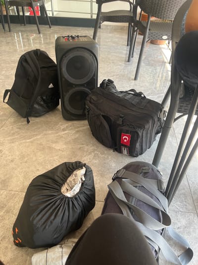 The scene depicts a tiled floor with four black bags and a speaker. The speaker, with two large speakers, is on the floor in the center of the image. The bags are spread out around the speaker, and one of the bags has a Turkish flag patch on it. The scene is located at latitude 38.198 and longitude 27.239 in the city of Gazi Paşa, Türkiye. The image appears to be taken from above, possibly from a nearby chair. The left side of the image includes a person's leg and foot, but no other individuals are visible. The floor is a light-colored tile, and the bags appear to be well-worn. The objects in the image are spread out and appear to be arranged randomly.