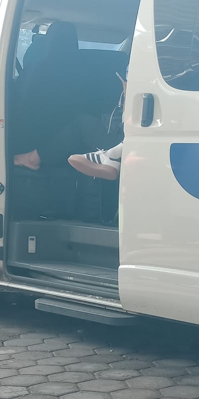 A white passenger van with its side sliding door open is parked on a grey hexagonal paver surface in the city of Bojongherang, Indonesia. Inside the van, two individuals are partially visible. The person closer to the open door has their right foot, clad in a white athletic shoe with black stripes and a brown gum sole, extended forward near the doorway. This individual's left hand, with a light skin tone, is seen near their head, potentially holding an object. A second individual is seated further inside the van, largely obscured by shadow, with only their bare left foot visible on the vehicle's floor. Charging cables or wires are present near the first individual's foot. A white rectangular object is affixed to the interior side panel of the van. The scene is illuminated by natural daylight.