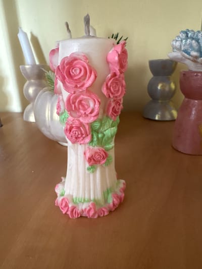 A close-up, eye-level shot shows a decorated white candle with pink roses and green leaves as accents. The candle is on a wooden surface, likely a table or shelf. In the background, out of focus, are several other decorative items, including a white candle in a holder, a white pear-shaped object, a grey hourglass-shaped object, and a pinkish-red object with a blue, sparkly top. The wall in the background is a pale yellow. The lighting appears to be natural and soft, coming from the right side of the frame. No people are visible. The scene is indoors.