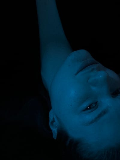 A person's face and neck are displayed in a stark blue hue against a dark background. The face is angled slightly, with one eye partially visible. The person's skin has visible pores and subtle shadows. The neck extends upward, disappearing into the darkness above. The overall image appears to be taken in low light, giving it a moody, ethereal quality. The subject's expression is neutral.