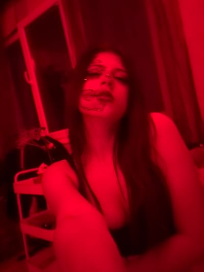 The image is a blurry, close-up, low-angle shot of a person illuminated by red lighting. The person has long, dark hair and appears to be wearing makeup. Their face is partially obscured by a jeweled or glittery mask-like decoration across their eyes and nose. The background is dark and indistinct, with what appear to be curtains or drapery and a window with a red frame visible on the left. A white rolling cart with shelves is partially visible in the lower left quadrant of the image. The scene does not provide enough information to determine the specific location in Aidemir, Bulgaria, or any specific actions or interactions.