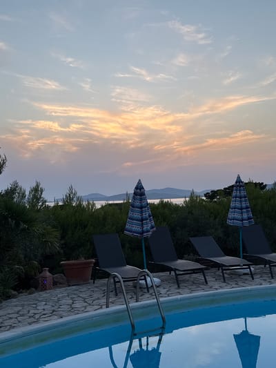 The image depicts a serene outdoor scene at dusk, likely from a private villa or resort in the city of Alghero, Italy. In the foreground, a clear blue swimming pool reflects the twilight sky. A metal ladder extends into the water from the pool's edge. Alongside the pool, a stone patio is furnished with several black lounge chairs and two patterned beach umbrellas, one blue and white, the other similar in design. Behind the patio, lush green foliage forms a dense hedge, with glimpses of palm trees and other vegetation. In the distance, a body of water separates the foreground from a hazy silhouette of hills or islands under a sky painted with soft hues of orange, pink, and pale blue, characteristic of a sunset. A decorative lantern with a blue light sits on the patio near a terra cotta pot. The overall atmosphere is tranquil and suggestive of a peaceful evening.