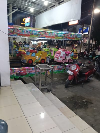This image depicts a nighttime scene in Sumberejo, Indonesia, featuring a brightly lit outdoor children's amusement ride named "Kiddy Ceria." The ride consists of multiple colorful vehicles, including a yellow truck, a green train car, and a pink train-like vehicle, positioned on a track or carousel mechanism. Several children, and at least one adult, are visible on or around the ride. The ride is covered by a canopy adorned with festive lights and banners displaying cartoon characters and the "Kiddy Ceria" logo.

In the immediate foreground, a series of light-colored tiled steps ascend from left to right. To the right of these steps and in front of the ride, a stainless steel stand with a rectangular basin is positioned. Beyond the ride, the ground is a dark, uneven surface, likely asphalt or concrete, where multiple motorbikes, primarily scooters of various colors (including red and light blue), are parked.

In the background, under overhead white canvas awnings or tarps, streetlights illuminate a commercial street. Visible signage includes a "Bank BRI" logo on a building and a blue sign with "m-din atm" text. Figures of people, mostly adults, are present in the distant background, suggesting general public activity. The overall environment indicates an urban setting, possibly a market or commercial area, during evening hours.