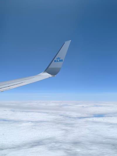 A view from an airplane window shows a KLM airplane wing against a backdrop of blue sky and a blanket of clouds. The KLM logo is visible on the wing. The location is not in Fischamend, Austria.