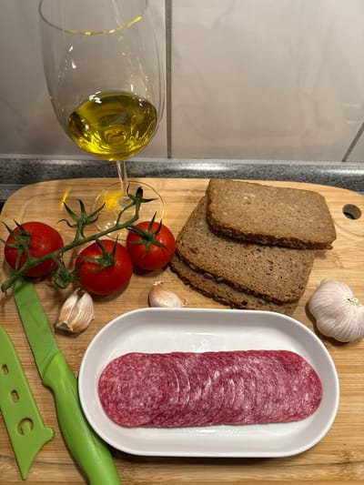 The image shows a wooden cutting board with ingredients for a meal. In the upper left corner is a wine glass filled with olive oil. To the right of the glass are three tomatoes on a vine. In front of the tomatoes are two cloves of garlic. To the right of the tomatoes and garlic are three slices of dark bread. In the bottom center of the image, a white oval dish contains several slices of salami. Two green-handled knives are also visible on the cutting board, one with its protective sheath. The background consists of a tiled wall and a countertop. The scene appears to be a kitchen counter. There are no people visible in the image, and no specific actions are being taken. The overall lighting suggests an indoor setting. The city of Velbert, Germany is not depicted in this image, and there is no indication of its location within this scene.