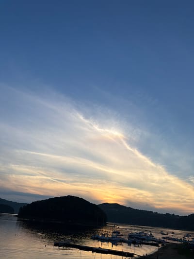 This image captures a scenic view of the Gródek Lake in Gródek nad Dunajcem, Poland, during what appears to be sunset. The sky is a gradient of deep blue at the top, transitioning to lighter shades of blue and then into warm hues of orange and yellow as it nears the horizon, painted with wispy clouds. Dominating the lower half of the image is the calm surface of the lake, reflecting the colors of the sky and the silhouettes of the surrounding landscape.

On the left, a densely forested island with a rounded top juts out into the water, its dark silhouette contrasting with the illuminated sky. The shoreline on the right is lined with numerous docked boats of various sizes, including sailboats and smaller motorboats, suggesting a popular recreational area. A wooden pier extends into the water from the right bank, with a few small figures visible on or near it, indicating human presence. The water itself is tranquil, with subtle ripples and reflections of the boats and the land. The overall impression is one of peacefulness and natural beauty at the close of day.