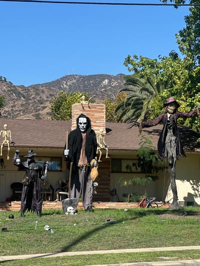 The image displays an outdoor daytime scene in Glendale, California, United States, featuring elaborate Halloween decorations on the front lawn and roof of a single-story residential property. Three prominent figures are displayed in the foreground: a central, approximately 12-foot tall grim reaper figure with a skull-like mask, a black hooded robe, brown tunic, and gray pants, holding a long shovel embedded vertically in the grass and a burlap sack featuring attached small skulls; to its left, a human-sized plague doctor figure in a black coat and beak mask, holding a lantern and a staff crowned with a skeleton hand; and to its right, an approximately 12-foot tall scarecrow figure with a tattered dark coat, purple-brimmed hat, and a grinning, toothy face, with outstretched arms.
Additional skeletal figures are displayed: one seated on the roof to the left, and two on a brick chimney, one standing and one seated. Small white skulls and dark spherical objects are scattered across the green grass lawn. The single-story house features a brown shingled roof, light-colored siding, and a visible address number '51' adjacent to the porch. Arid, brush-covered hills with communication towers on their peak rise in the background under a clear blue sky. A large palm tree and other leafy green trees are visible around and behind the house. Shadows cast by the figures and trees indicate bright sunlight.