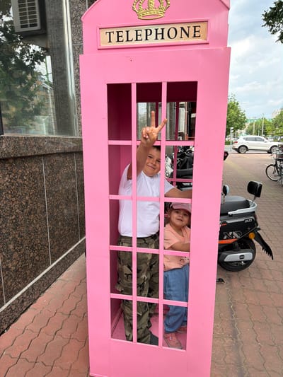 The image shows two young children inside a bright pink, British-style telephone booth. The booth has a sign at the top that reads "TELEPHONE" in gold lettering on a cream background, topped with a golden crown. The larger child, a boy, stands in the upper section of the booth, wearing a white t-shirt and camouflage cargo pants, and is making a peace sign with his right hand. The younger child, a girl, is in the lower section, wearing a light orange t-shirt and blue pants, and is smiling at the camera. The telephone booth is positioned on a red brick sidewalk. To the right of the booth, there is a black and silver electric scooter. In the background, there are trees, buildings, and a white SUV. The scene appears to be outdoors in an urban environment.