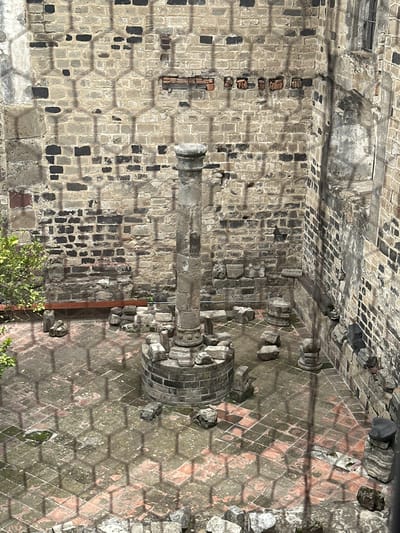 The image shows a courtyard at the Ex-Convento de San Miguel Arcángel in Huejotzingo, Mexico. A large stone column stands in the center of the courtyard, surrounded by a circular base constructed from bricks. Scattered around the courtyard are various stone fragments, some appearing to be architectural remnants. The walls of the courtyard are made of stone and brick, showing signs of age and weathering. The ground is paved with reddish-brown tiles, some of which are covered in moss or debris. A small green bush is visible on the left side of the frame, near the wall. A faint hexagonal pattern, likely a shadow from a fence or grate, is superimposed over the scene. There are no people visible in the image. The overall impression is one of an ancient, possibly archaeological, site within the historic convent.