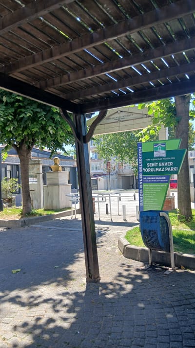 The image depicts an outdoor scene, likely a park or public space, in the Bahçelievler district of Turkey. The foreground is dominated by a wooden pergola structure casting dappled shadows on a paved walkway. To the right, a green sign with white text reads "BAHÇELİEVLER BELEDİYESİ" (Bahçelievler Municipality) and "ŞEHİT ENVER YORULMAZ PARKI" (Martyr Enver Yorulmaz Park), indicating the park's name and location. Below this, it lists "DR. HAKAN BAHADIR BAHÇELİEVLER BELEDİYE BAŞKANI" (Dr. Hakan Bahadır, Mayor of Bahçelievler). Further text on the sign includes a website address and bullet points detailing park regulations or features, such as "Halkın huzur ve rahatı için temiz tutulmasından sorumludur" (Responsible for keeping it clean for the peace and comfort of the public) and "Evcil hayvanlarınızın dışkılarının alanda bırakılmaması" (Pet droppings not to be left in the area). A blue trash receptacle is positioned near the sign.
In the mid-ground, a tree with green foliage stands to the left, next to a marble monument topped with a golden bust. Behind the monument, buildings with multiple windows and balconies are visible. The walkway is paved with dark grey cobblestones. White railings and tables suggest an area for seating or recreation. The environment appears to be sunny, with bright light illuminating the scene and creating distinct shadows. The overall impression is one of a well-maintained public park.