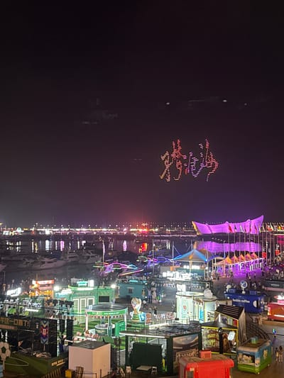 The image depicts a nighttime scene at the Shinan District, China, specifically along a waterfront area that appears to be a vibrant entertainment or festival zone. In the sky, a drone light show forms Chinese characters, with vibrant pink and yellow dots creating illuminated calligraphy against the dark night sky. Below, a harbor is filled with numerous yachts and boats, their lights reflecting on the water. Along the shoreline, there is a lively pedestrian area with various stalls, tents, and structures, many of which are brightly lit with colorful LED lights in shades of pink, purple, blue, and green. Some stalls have signage, including one with "FIFA WORLD CUP" branding and a trophy. People are visible walking around, congregating, and interacting within these illuminated areas. The overall atmosphere suggests a bustling night market, festival, or cultural event by the sea.