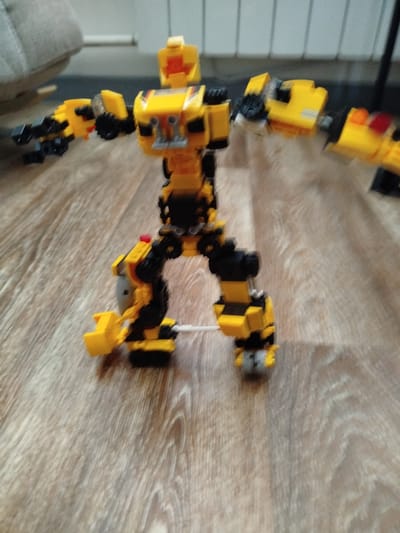 This media file shows a yellow and black Lego transformer figure standing on a wooden floor. The figure is built from what appear to be Lego Technic pieces and has a robot-like appearance with arms and legs. Its body is primarily yellow with black accents and some red and orange pieces visible, likely representing lights or details. The figure's legs are spread apart in a stance, and its arms are positioned outward. One arm appears to have a claw-like attachment, and the other is bent at the elbow. A thin white wire or string is visible extending from the figure's left leg. The background is blurred but suggests an indoor setting with furniture and vertical blinds or a wall. The lighting indicates it is daytime. There is no visible text in the image. The scene is static, with the Lego figure posed on the floor.