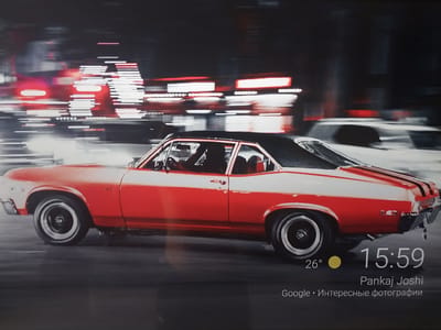 The image shows a red and white classic muscle car driving at night on a blurred city street. The background is filled with streaks of red and white lights, suggesting motion blur from passing vehicles and illuminated buildings. In the lower right corner, a digital display shows the time as 15:59, a temperature of 26 degrees, and the text "Pankaj Joshi" followed by "Google • Интересные фотографии". The context of Nizhneye Myachkovo, Russia is not discernible from the visual information provided.