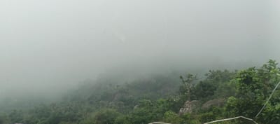 This media file captures a view of a lush, green, hilly landscape in Gidan Waya, Nigeria, almost entirely engulfed in dense fog or mist. The upper half of the image is dominated by the thick, white-grey mist, severely limiting visibility of the horizon or sky. In the upper central area of the mist, a faint, circular, halo-like reflection or lens flare is discernible.

Below the heavy mist, the terrain is covered with abundant green trees and shrubs, indicative of a forested or vegetated hillside. Scattered among the foliage are large, grey rocks. The lower-right corner of the image shows a metal railing or wire, suggesting the viewpoint is from an elevated position or a structured overlook.

Notable details include graffiti text on two visible rocks in the middle-right section of the image. One rock faintly displays the letters "DVD". Below it and slightly to the right, another rock has "SVC" written above the numbers "6170". The weather is overcast and very misty. No people or specific activities are visible in the scene. The overall impression is one of natural beauty shrouded in a serene, mysterious fog.