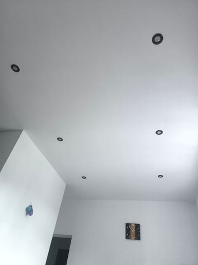 The image depicts a modern, interior setting, likely a home or office space in Katowice, Poland. The scene is dominated by a white, textured ceiling with multiple recessed, black-rimmed spotlights. One spotlight is visible in the upper left quadrant, another in the upper right, and three more are spread across the mid-section of the ceiling.
On the wall, in the lower right portion of the image, a square clock with a dark face and wooden accents is mounted. The clock features Arabic numerals and has black hands pointing to approximately 10:09. To the left, on another wall, two small, colorful objects, appearing to be balloons or similar inflatable items, are attached to the wall with a hook. They are a light blue and a purple color.
The overall lighting suggests it is daytime, with light entering from an unseen source, casting soft shadows. The environment appears clean and minimalist, with a focus on architectural features like the angled walls and ceiling design. There are no people visible in the image. The prominent objects are the ceiling lights, the wall clock, and the decorative wall items. The setting is calm and uncluttered.