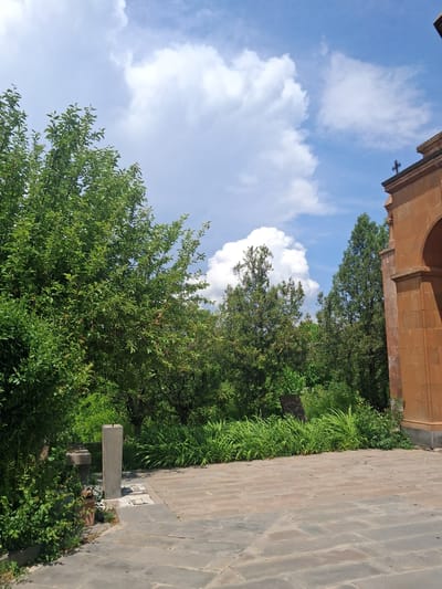 The image shows a sunny day with a partly cloudy sky. The scene is outdoors and appears to be in a garden or park area next to a building with a terracotta-colored facade. The building has an arched opening and a small cross visible on its upper edge. A paved walkway made of large, grey flagstones runs along the right side of the image, bordering lush green vegetation. The vegetation includes trees with dense foliage and tall, grass-like plants. A low, grey stone bench is situated in the foreground, next to the paved walkway and amidst the greenery. The image is taken from a low angle, looking upwards towards the sky and the building. The overall impression is one of a tranquil outdoor setting, possibly a historical or religious site given the cross on the building. The lighting suggests it is daytime, likely late morning or afternoon, with bright sunlight illuminating the scene. The weather appears pleasant. The location is identified as Martiros, Armenia.