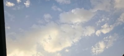 The image shows a sky filled with scattered clouds, predominantly white and light gray, against a pale blue background. The light suggests it could be late afternoon or early morning. On the left edge of the image, a dark, angled structure, possibly part of a building or roofline, is visible. The scene is an outdoor environment, likely an urban setting in Margilan City, Uzbekistan, given the provided context. There are no people or discernible subjects visible in the frame, and no specific actions or interactions are depicted. The focus is solely on the atmospheric conditions.