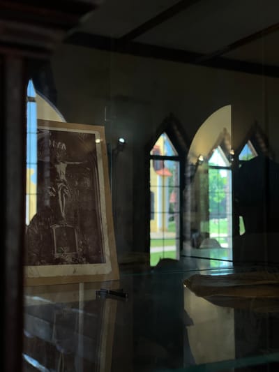 The image shows a sepia-toned photograph of the crucifixion of Jesus Christ, displayed inside a glass case. The photograph is old and appears to be mounted on cardboard. The background of the photograph depicts an ornate architectural setting. The glass case reflects the exterior, revealing arched windows with stained glass, through which a sunlit garden and building can be seen. The setting suggests an indoor display, possibly in a museum or historical site, located within the Літвенскі сельскі Савет, Belarus. The lighting is dim inside the display case, with highlights on the glass and the photograph. There are no people visible in the immediate foreground or interacting with the photograph. The overall scene is still and contemplative, focusing on the religious artifact.