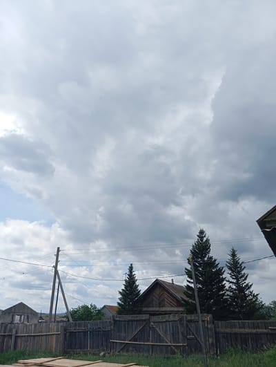 This is a vertical, eye-level, outdoor, full shot of a cloudy sky over a rural landscape in Novodesyatnikovo, Russia. The sky is filled with large, dramatic grey clouds, with patches of lighter blue sky peeking through in the upper left. In the middle ground, a wooden fence runs horizontally across the frame, with a large wooden gate in the center. Behind the fence, there are several houses, including a rustic wooden house with a dark roof, and a more modern grey concrete building. Three tall, dark green fir trees are visible behind the fence and to the right of the wooden house, partially obscuring it. A wooden utility pole with wires stands to the left of the trees. In the foreground, there are several light-colored wooden planks laid out on the grass. The overall impression is of a quiet, overcast day in a rural village. The weather appears to be cloudy, possibly with a chance of rain. The time of day is difficult to determine precisely due to the overcast conditions, but it appears to be daytime. There is no visible text in the image.