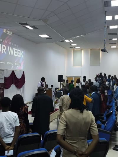 This image depicts an indoor gathering in Kızılbaş, Northern Cyprus / Cyprus. The scene shows a spacious room with white walls and a white drop-tile ceiling fitted with numerous square fluorescent light fixtures. On the left, a large projection screen displays text including "AFFIRMATIONS FOR OUR WEEK" and "MANIFESTING THE KINGDOM OF GOD," along with social media handles such as "@rnecfellowship" and "@rnec.church." Below the screen, a stage area is visible with a white backdrop draped with burgundy fabric.
At the front of the stage, a male speaker in a white shirt and burgundy vest stands behind a podium, holding a microphone and addressing an audience. To his right, a large black speaker is positioned. Further right and behind the speaker, several individuals are visible, including musicians with drums and other instruments, suggesting a live band. A white stand adorned with flowers is also present on the stage.
A crowd of people, predominantly adults, fills the room, most standing and facing the stage. The individuals are dressed in varied attire, ranging from casual to semi-formal. In the foreground, viewed from behind, are several audience members: a person in a white t-shirt holding a small white object, a person with dark curly hair and a patterned dress, a man in a black tunic, and a woman in a tan dress with braided hair. Rows of empty and occupied blue upholstered seats with black frames are visible in the foreground and extending into the middle ground. On the right wall, two large rectangular windows covered by light-colored roller blinds are present. A projector is mounted from the ceiling on the right side of the room.