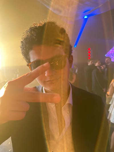 The image depicts an indoor event scene in the city of Huizúcar, El Salvador. In the foreground, a male individual with curly dark hair is centrally positioned, looking towards the camera. He wears brown-framed sunglasses, a white collared shirt with the top buttons open, and a dark suit jacket. A small, dark stud is visible on his lower lip. His left hand is raised directly in front of him, with the index and pinky fingers extended and the middle and ring fingers bent, forming a gesture. The scene is affected by a prominent warm-toned lens flare originating from the left, casting a glow across the subject and the left side of the frame. The background is dark and out of focus, populated by several indistinct figures, some appearing to be in formal or semi-formal attire, suggesting a social gathering. A distinct blue light beam projects diagonally downwards from the upper right, and a vertical series of four red circular lights is visible in the mid-right background. To the far right, a multicolored, possibly geometric, light fixture is partially visible.