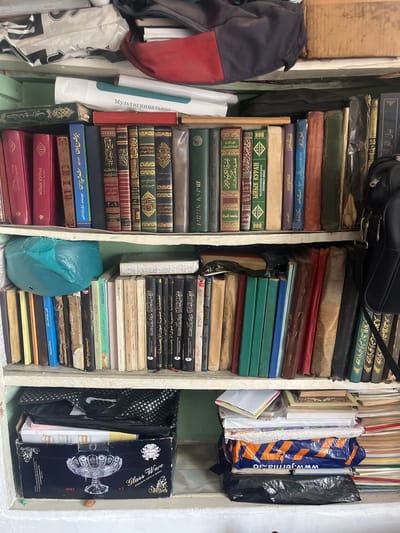 The image displays a shelf filled with books and other items. On the top shelf, there are several books, some with red and green covers, and a blue bag. A red and blue bag is also visible on the top of the shelf. The middle shelf is packed with numerous books of various sizes and colors, including some with Arabic script. There is also a blue plastic bag and a white book. The bottom shelf contains a dark box with "Glass Ware" written on it, along with some folded items and a black plastic bag. The overall environment appears to be a storage area or a room with shelves. The scene is set in the city of Tashlak, Kyrgyzstan.
