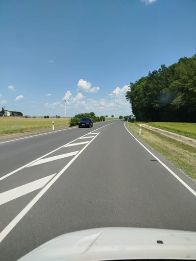 A black SUV is driving on a two-lane asphalt road on a sunny day. The road has white lane markings. The SUV is in the right lane, heading away from the camera. To the left of the road, there is a green field with some white fences and buildings in the distance, as well as a wind turbine. To the right of the road, there is a dense green forest. The sky is blue with some scattered white clouds. The view appears to be from the perspective of the driver of another vehicle, with the hood of a white car visible in the foreground.