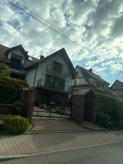 The image shows a street view of several detached houses in Wilkowisko, Poland. The houses are multi-story with red-tiled roofs and white facades. Some houses have balconies with black wrought-iron railings. There is a paved driveway leading to a black metal gate, which is partially open. Lush green bushes and trees surround the houses, and a neatly paved sidewalk runs along the street. The sky is cloudy with patches of blue sky visible. There are power lines running across the top of the image. No people are visible in the image.
