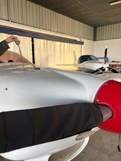 The image depicts an interior view of an aircraft hangar at the Aérodrome de Toussus-le-Noble, France. In the foreground, the front section of a white single-engine propeller aircraft is visible. A human hand, clad in a dark green sleeve, is positioned on the left, holding a metal dipstick inserted into a fuel port on the aircraft's fuselage, indicating a fuel level check. The aircraft features a prominent red propeller spinner, and one propeller blade is partially covered by a black fabric sleeve. Below the spinner, mesh-covered air intakes are present on the cowling. In the blurred background, a second white single-engine aircraft with a dark canopy and propeller is parked further inside the hangar. The hangar's walls and ceiling are constructed of corrugated metal, and a large, light-colored curtain or tarp covers a hangar door opening in the rear, providing ambient illumination.
