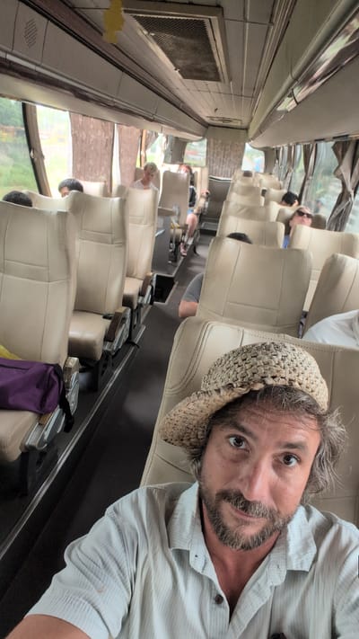 The image depicts the interior of a passenger bus, likely in Somsavan, Laos, seen from a low-angle perspective. The foreground is dominated by the face of a man wearing a straw hat and a light-colored striped shirt. His gaze is directed towards the camera, and he has a beard and medium-length hair. The background reveals rows of beige upholstered bus seats stretching towards the front of the vehicle. Several other passengers are visible seated in the bus, some facing forward and others looking away from the camera. A purple bag is on a seat in the left foreground, and a yellow air freshener hangs from the ceiling. The windows on the sides of the bus show a blurred view of greenery and some structures, suggesting an outdoor environment. The lighting appears to be natural, coming from the windows and the interior lights of the bus.