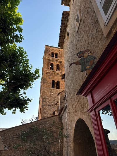 The image depicts a narrow street scene in Moustiers-Sainte-Marie, France, with a tall, ancient stone bell tower dominating the background. In the foreground, a weathered stone building occupies the right side, featuring an arched entryway and a prominent red-trimmed window. A painted wooden sign of a jester figure is affixed to the building's facade. Inscriptions are visible on the red trim of the window. A large tree with green leaves is situated to the left, partially obscuring the view of the bell tower and a portion of the sky. The overall scene suggests a historic, possibly medieval, European town.