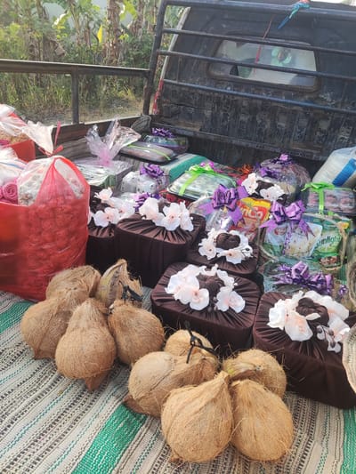 In Ngentrong, Indonesia, a pickup truck bed is filled with gifts and coconuts. The gifts are primarily food items and are wrapped in clear plastic with purple ribbon accents. Some of the gifts appear to be boxed sweets or snacks, while others are larger, possibly cakes or larger food arrangements. Several brown fabric-covered boxes are also present, each adorned with white artificial flowers. In front of the gifts, a pile of coconuts, still in their husks, rests on a woven mat. A large red plastic bag filled with what appears to be more red flowers or flower garlands is also visible. The background shows lush green foliage and trees, indicating a rural or semi-rural setting. The truck itself appears weathered, with a black metal frame around the bed and a cracked rear window. The overall scene suggests a preparation or delivery of items for an event, possibly a wedding or celebration, in the village of Ngentrong.