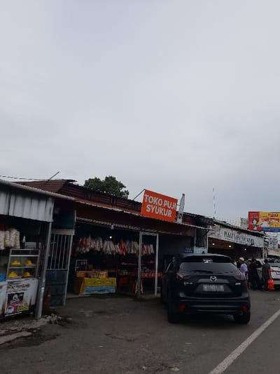 The image shows a street scene in Kecamatan Purwoasri, Indonesia. A black car is parked along the side of the road, facing away from the camera. The vehicle is located in front of a store with a sign that reads "TOKO PUI SYUKUR". The store has a canopy over the storefront and shelves lined with various items. There is a small tree growing in the background. There are three people visible in the scene, walking past the parked car. The sky is clear and overcast. The road is paved and there is a white line down the center. There are a few other cars in the distance.