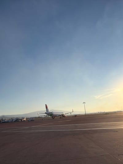 An airplane is parked on an airport tarmac under a hazy, bright sky. The aircraft is positioned alongside a service road and appears to be ready for boarding or departure. The tarmac stretches into the distance, meeting a row of support vehicles and infrastructure. A light pole stands nearby. The image is oriented on its side.