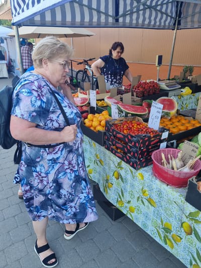 The image depicts an outdoor market scene in Kuršėnai, Lithuania. A woman wearing a floral dress and black sandals is standing at a fruit and vegetable stall, looking at a carton of cherry tomatoes. She has blonde hair and is wearing glasses. To her right, a second woman in a dark patterned blouse is standing behind the stall, seemingly attending to customers or arranging produce. The stall is laden with various fresh produce, including watermelons, peaches, tomatoes, strawberries, pineapples, and garlic. Price tags are visible on some of the items. The stall is covered by a striped blue and white awning. In the background, a bicycle is visible, and a building with an orange wall can be seen. The ground is paved with grey bricks. The overall scene is characteristic of a local farmers' market.