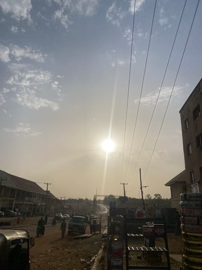 The image depicts a street scene in Unguwar Fatika, Nigeria. The sun is high in the sky, creating a bright glare. Power lines run across the upper left. Buildings line the street, including multi-story structures and smaller shops. Vehicles and pedestrians are visible on the street. A rack filled with tires and motor oil products sits in front of one of the buildings.
