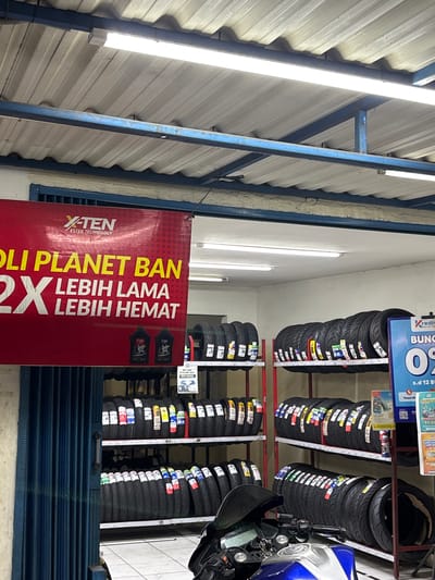 The image shows the interior of a tire shop, likely located in Binong, Indonesia. The shop features rows of motorcycle tires displayed on metal shelves. A red banner hangs prominently, advertising "X-TEN ESTER TECHNOLOGY" and a promotion stating "2X LEBIH LAMA LEBIH HEMAT" (2X Longer Lasting, More Economical). Two bottles of oil are visible on the shelf below the banner. To the right, a blue promotional poster displays "KREDIT", "BUNGA 0%", and text indicating a sale from "s.d 12 B". A motorcycle is partially visible in the foreground, suggesting it's a customer's or a display item. The shop appears to be well-lit with fluorescent lighting from the ceiling. The overall environment is that of a commercial establishment focused on automotive parts, specifically tires.
