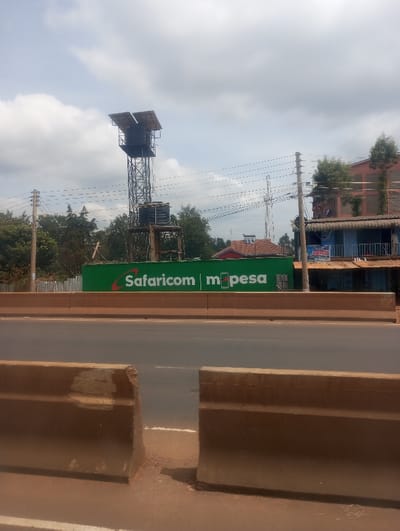 The scene depicts an outdoor, daytime view in Gitaru ward, Kenya, under an overcast sky. In the immediate foreground, two weathered, earth-toned concrete road barriers, showing visible fracturing at their upper edges, flank a dirt median that separates them from a paved road. Beyond these barriers, a multi-lane asphalt road surface is visible, featuring faint white lane markings.
Across the asphalt road, a large green retaining wall or barrier is prominently displayed, bearing the white and orange "Safaricom m-pesa" logo. Behind this green structure, a tall, metallic lattice tower stands, supporting a flat array of dark solar panels at its peak and two black water storage tanks mounted lower on its frame. To the left of the tower, dense green tree foliage is present. To the right, multiple wooden utility poles with overhead electrical lines extend across the midground.
The background includes a low-rise building with a distinct red-tiled roof, a slender, more distant lattice communication tower, and a multi-story building with a reddish-orange facade and a lower blue section, partially obscured by green trees. No human figures, vehicles, or dynamic actions are discernible within the frame.