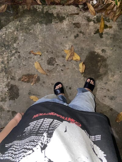 A first-person perspective image shows a person standing on a wet, concrete surface covered in fallen leaves. The person is wearing black slides, blue jeans, and a black t-shirt with white and red text and graphics. The t-shirt has the word "CONTRADICTION" printed in red across the chest. A blurred portion of the person's arm is visible on the left side of the frame. The environment appears to be outdoors, possibly a sidewalk or pathway, with scattered brown and yellow leaves. The concrete shows signs of dampness and some staining. No specific landmarks indicating Banjarmasin, Indonesia are visible.