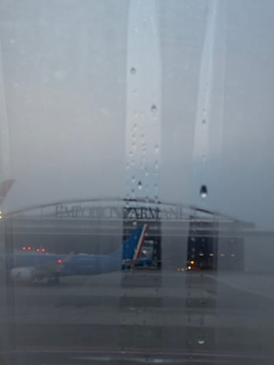 The image shows a view through a rain-spattered window, partially obscuring a building. The words "EMPORIO ARMANI" are visible vertically on the building's facade. There is a flag visible, featuring red, white, and green stripes (potentially Italian). A string of small, red lights is visible, as well as yellow lights. The location is likely Peschiera Borromeo, Italy, due to the presence of Emporio Armani and the Italian flag.