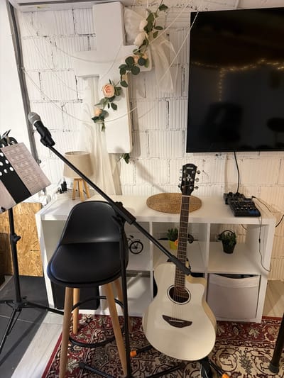 This image captures an indoor setting, likely a home music studio or practice area, characterized by musical equipment and decorative elements. A white Yamaha acoustic guitar, identifiable by the "YAMAHA" text on its headstock, leans against a white shelving unit on the right side of the frame. This unit holds various items including small potted plants and a woven mat. An audio mixer is mounted on the wall above the shelving.

To the left, a large black flat-screen television is mounted on a white, textured wall, reflecting the glow of fairy lights. Below the television, a prominent white decorative cross, adorned with artificial pink roses, green leaves, and white sheer fabric, hangs on the wall. Pearl-like beaded strings are draped across the cross and extend into the foreground.

In the foreground, a microphone on a black stand is positioned in front of a sheet music stand, which holds visible sheet music. Behind the microphone stand is a black chair with wooden legs. A small table lamp with a beige shade is also visible. The floor features a section of a patterned red and white rug and an area covered by oriented strand board (OSB). Cables are visible, connecting the electronic devices. No people are present, and the scene suggests a setup ready for music or a quiet resting place for instruments.