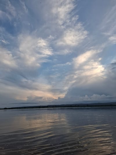 The image depicts a wide, calm river reflecting a dramatic sky filled with clouds. The sky transitions from a pale blue at the top to darker, textured clouds towards the horizon. Some clouds are illuminated by sunlight, creating a warm glow, while others appear darker and more ominous. The river occupies the lower half of the frame, its surface disturbed by gentle ripples. A dark, silhouetted line of trees and hills forms the distant shore. The scene appears to be set in the natural environment surrounding Малая Сазанка, Russia, likely during a time of day with dynamic atmospheric conditions. There are no people or specific man-made objects visible in the image.