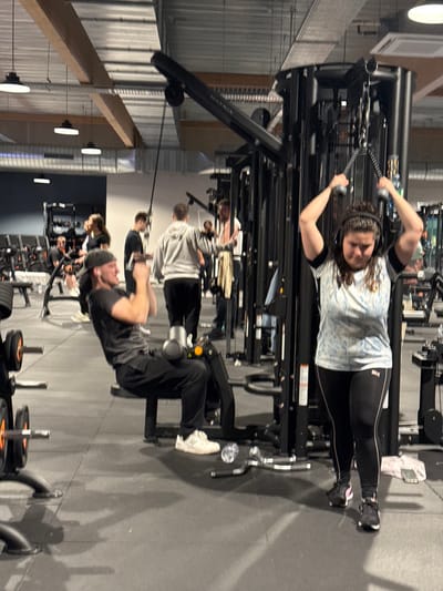 In a gym setting, a woman wearing a blue patterned shirt and black leggings stands at a cable machine, pulling down on the handles. Several people are working out on various machines in the background, including a man performing crunches. The gym has a high ceiling with exposed beams and industrial lighting. Exercise equipment and weights are scattered throughout the space.