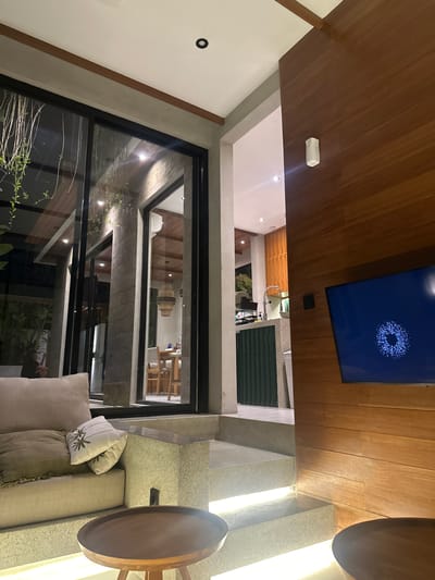 The image shows an interior space in Tumbak Bayuh, Indonesia. A large window offers a view of an outdoor area with plants. Inside, there's a seating area with a light-colored sofa and decorative pillows. A round wooden table stands nearby. The ceiling is constructed of wood planks, and a television screen is mounted on it.