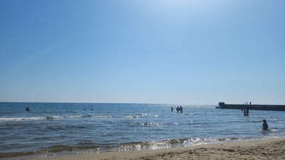 The image depicts a bright, sunny day at a beach in Sabaudia, Italy. The sky is a clear, cloudless blue, with a faint rainbow-like arc visible in the upper left quadrant, likely due to the sunlight interacting with atmospheric moisture. The sun is positioned high in the sky, casting a bright glare on the water's surface, creating numerous sparkling reflections.

The foreground shows a sandy beach with subtle ripples indicating where the water has recently receded. The sea is calm with gentle waves lapping at the shore. Several people are visible in the water. On the left side of the image, a lone figure stands in the water. Further to the right, a group of people are wading and playing in the sea. In the mid-ground, a concrete structure, possibly a pier or breakwater, extends from the right side of the frame into the sea. Silhouetted figures can be seen standing on this structure. On the far right, a person sits partially submerged in the water near the shore. The overall impression is a relaxed, summery atmosphere on the coast of Sabaudia.