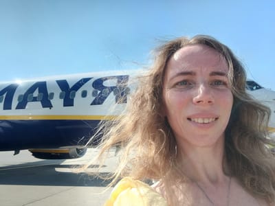 A woman is taking a selfie with a Ryanair airplane in the background. The airplane has the Ryanair logo on its side and is parked on the tarmac at Weeze Airport in Weeze, Germany. The woman has long, light brown hair that is blowing in the wind. She is smiling and wearing a necklace. The sky is clear and blue.