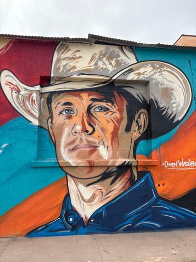 The image shows a mural of a man wearing a cowboy hat, painted on the side of a building in Lordsburg, New Mexico, United States of America. The mural features the man's face, rendered in vibrant colors with bold lines and dramatic shading. He has blue eyes and a serious expression. The background of the mural is abstract, with splashes of red, teal, and orange. The mural is located on a textured wall, and there is a small window in the center of the painting that has been covered with a grate. The signature "OWEN WASHBURNE" is visible in white spray paint on the right side of the mural, near the bottom. The building appears to have a corrugated metal roof. The overall impression is of a striking and colorful piece of public art.