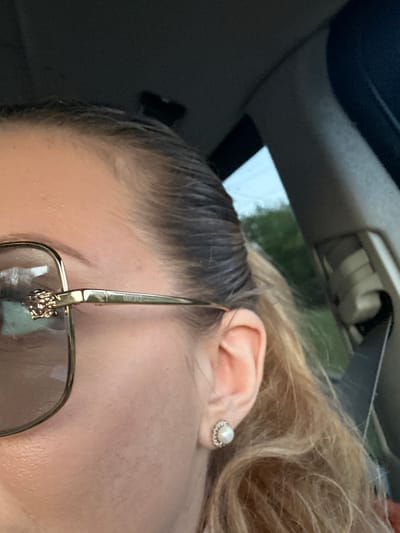 The image shows a close-up view of a person's face from the cheek up, partially obscured by sunglasses with gold frames. The person is wearing an earring in their right ear. The background includes elements of a vehicle interior, such as a sun visor and headliner. The location cannot be determined from the provided image.