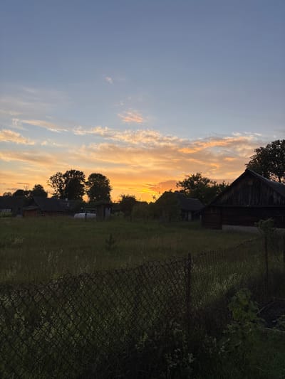 The image depicts a rural sunset scene in the Ганчарскі сельскі Савет, Belarus. A chain-link fence runs across the foreground, with tall, green grass and some wildflowers growing behind it. In the middle ground, a cluster of traditional wooden houses and outbuildings are visible, silhouetted against the vibrant orange and yellow hues of the sunset sky. Several mature trees with dark foliage are scattered amongst the buildings. The sky transitions from a clear, pale blue at the top to bands of illuminated clouds near the horizon, where the sun is setting. The overall impression is one of a quiet, rustic evening in the Belarusian countryside.